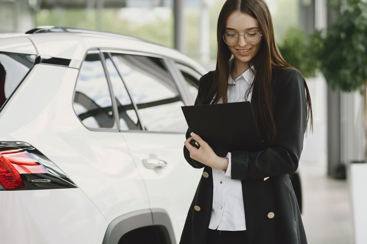 founder-img Smiling woman in a black jacket reviewing documents near a white car indoors.